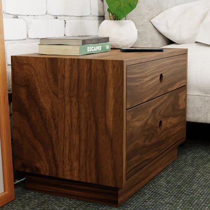 Beside a bed, the Krovel Freestanding Nightstand in Walnut showcases its handmade charm and two soft-close drawers. Books and a tablet adorn its top, set against a white brick wall, with parts of a gray sofa and green plant leaves enhancing the background.