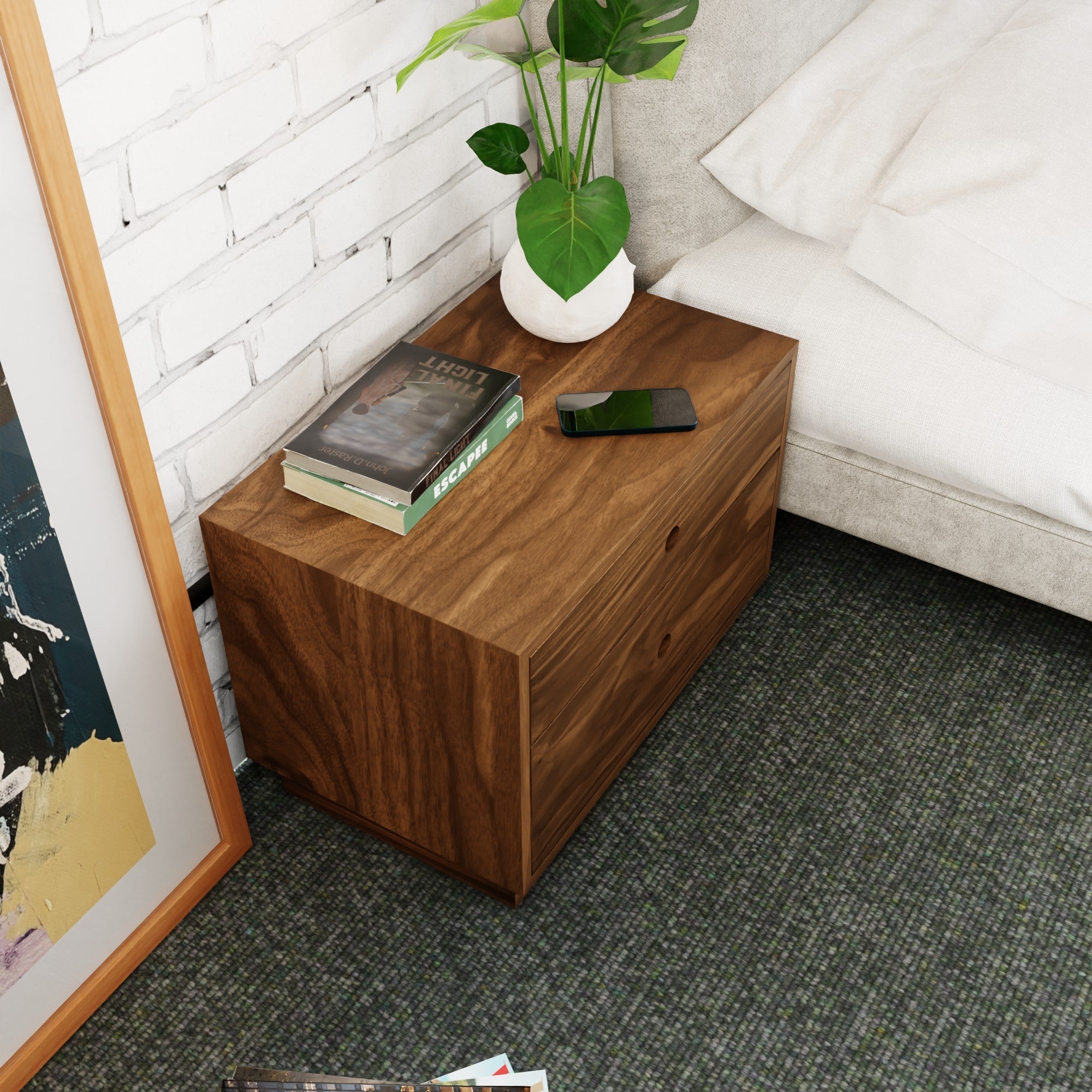 A Krovel Freestanding Nightstand in Walnut is next to a bed on carpet, featuring two soft-close drawers. On top are two books, a smartphone, and a potted plant with large green leaves. A framed picture leans against the white brick wall.