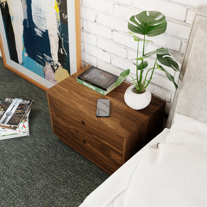 The Krovel Hardwood Freestanding Nightstand with two drawers holds a potted plant, phone, and book. An abstract painting leans against a white brick wall beside the nightstand next to a bed with white bedding. A magazine lies on the floor.