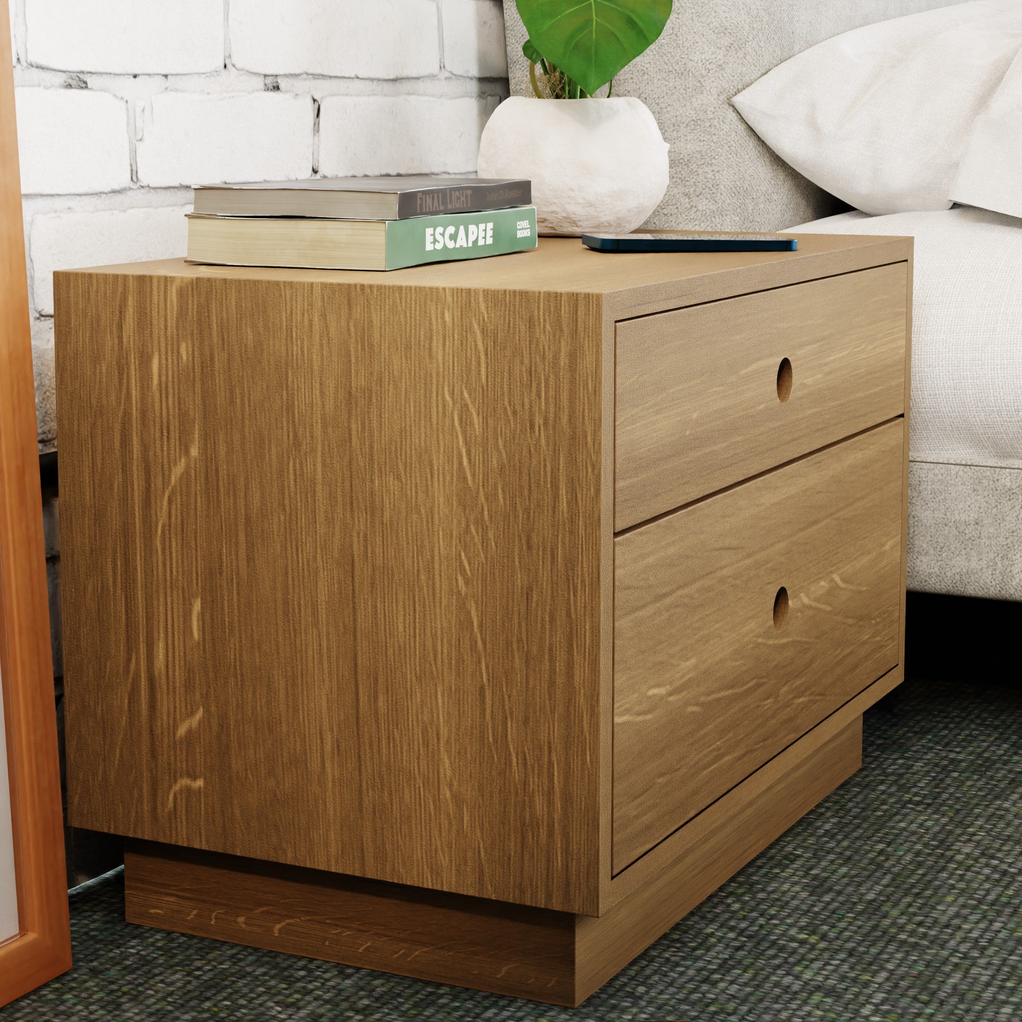 A Krovel Freestanding Nightstand in White Oak, crafted from solid wood, stands by the bed against a white brick wall. It features soft-close drawers and is topped with books like "Escape" and a closed laptop. A partially visible plant adds greenery in the background.
