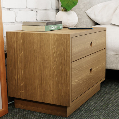 A Krovel Freestanding Nightstand in White Oak, crafted from solid wood, stands by the bed against a white brick wall. It features soft-close drawers and is topped with books like "Escape" and a closed laptop. A partially visible plant adds greenery in the background.