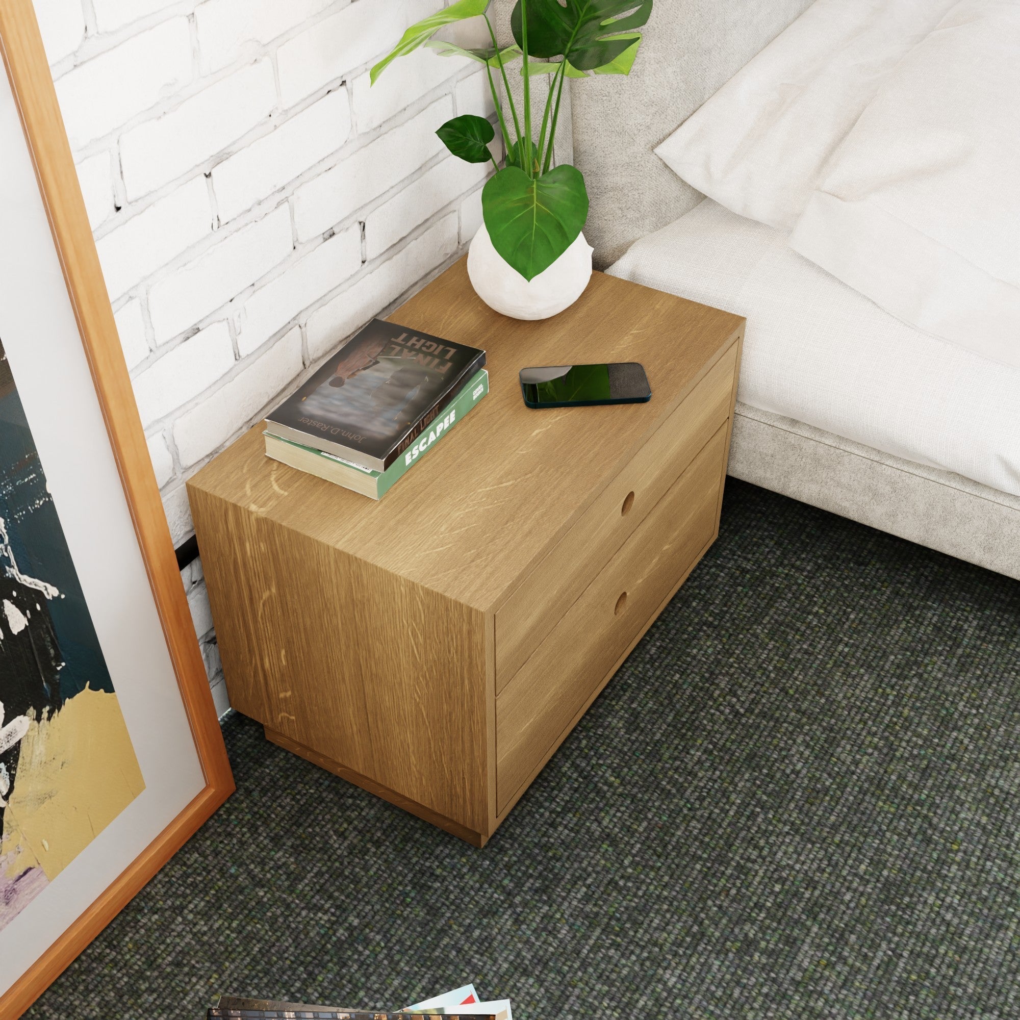 A cozy bedroom showcases a Krovel Freestanding Nightstand in solid White Oak beside the bed, holding books, a smartphone, and a small plant. The carpeted floor complements the setting, with a framed print leaning against the white brick wall.