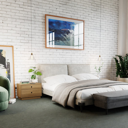 A modern bedroom includes a white brick wall, a double bed with gray bedding, and Krovel's Freestanding Nightstand in solid White Oak. Two hanging lights and green plants enhance warmth. Abstract art adorns the wall above the bed, complemented by a green chair and dark bench.