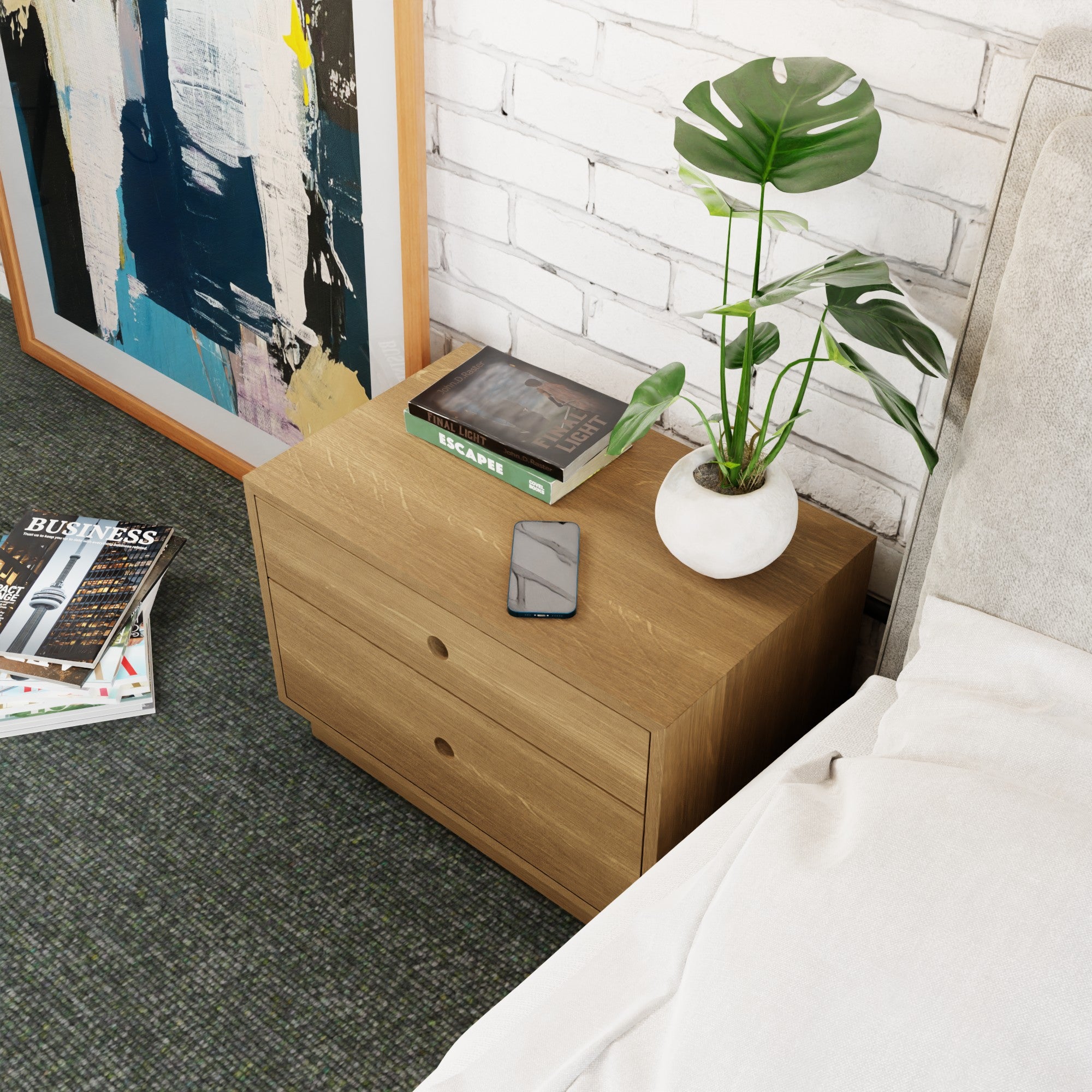 The Krovel Freestanding Nightstand in White Oak, with two soft-close drawers, complements a bed while holding a potted plant, smartphone, and magazines. An abstract painting leans against the white brick wall on the green carpeted floor.