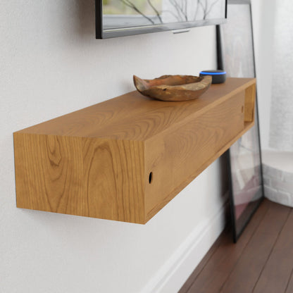 A Krovel Media Console Table in Cherry, finished with VOC-free polymerized linseed oil that highlights the rich grain, is styled under a wall-mounted TV. It's adorned with a decorative bowl and a small blue container. A framed picture leans against the wall on the dark wood floor near a window.