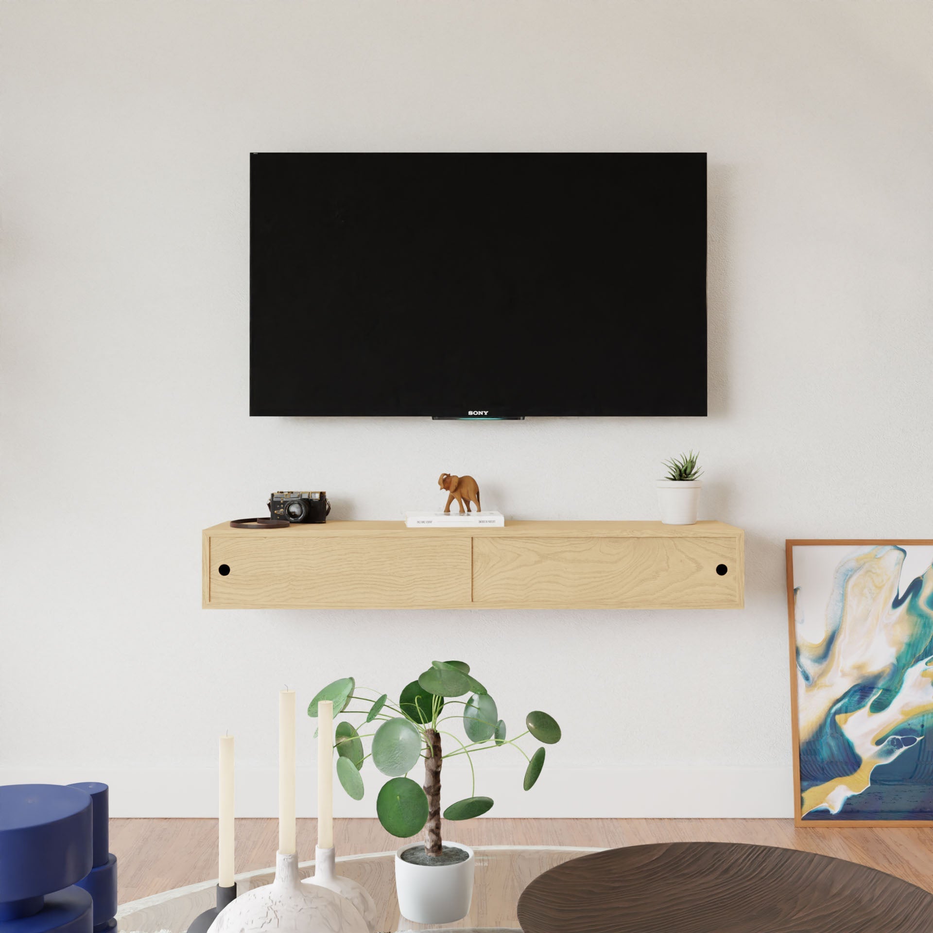 A minimalist living room features a wall-mounted TV above a Krovel Maple Media Console Table. The table displays a camera, an elephant figurine, and a small plant. In the foreground, candles and another plant adorn the American Cherry table, while abstract art decorates the walls.