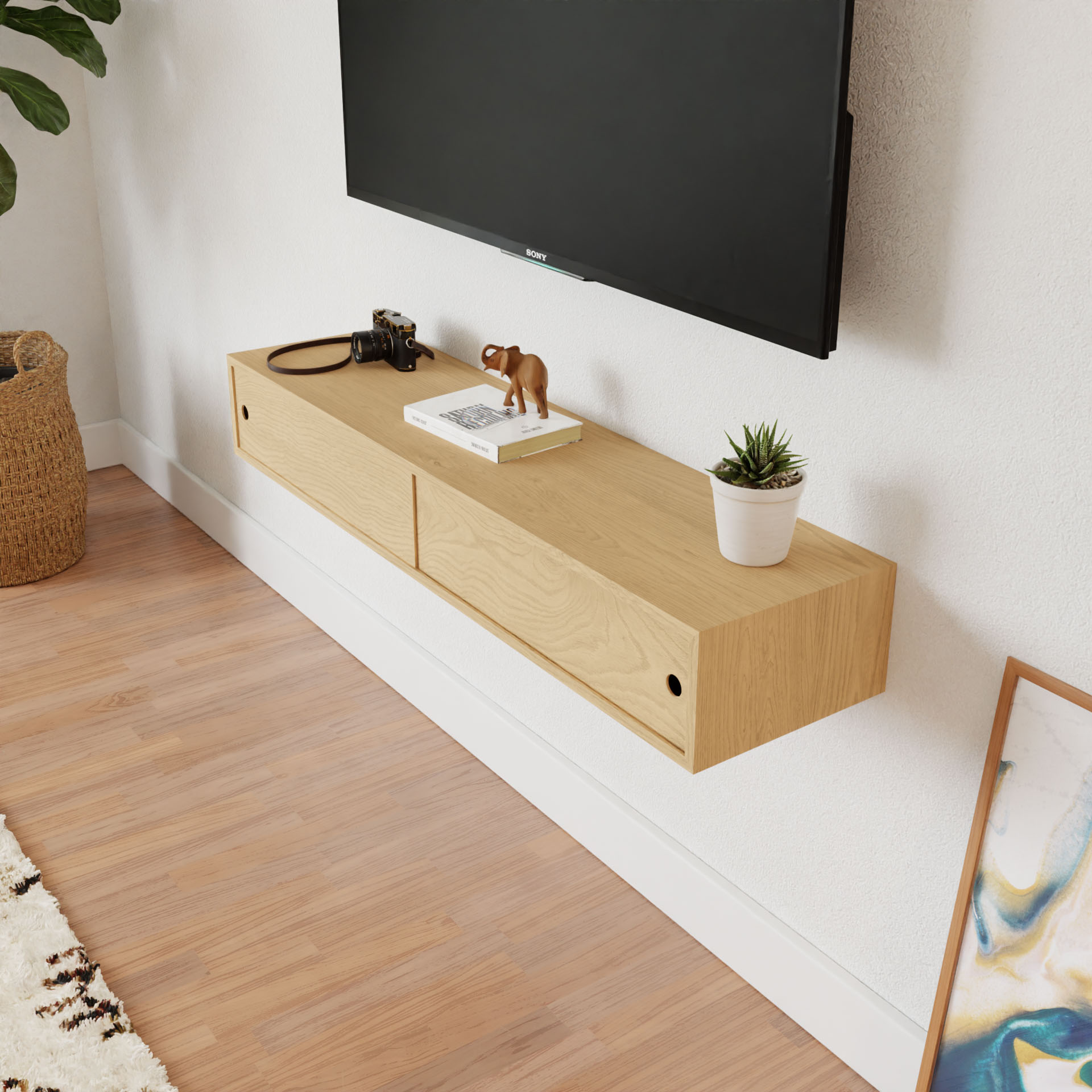 The Krovel Media Console Table in Maple, mounted on the wall beneath the flat-screen TV, showcases decor like a camera, small animal figurine, and potted plant. Framed artwork leans casually against the wooden floor with a plant basket behind it.