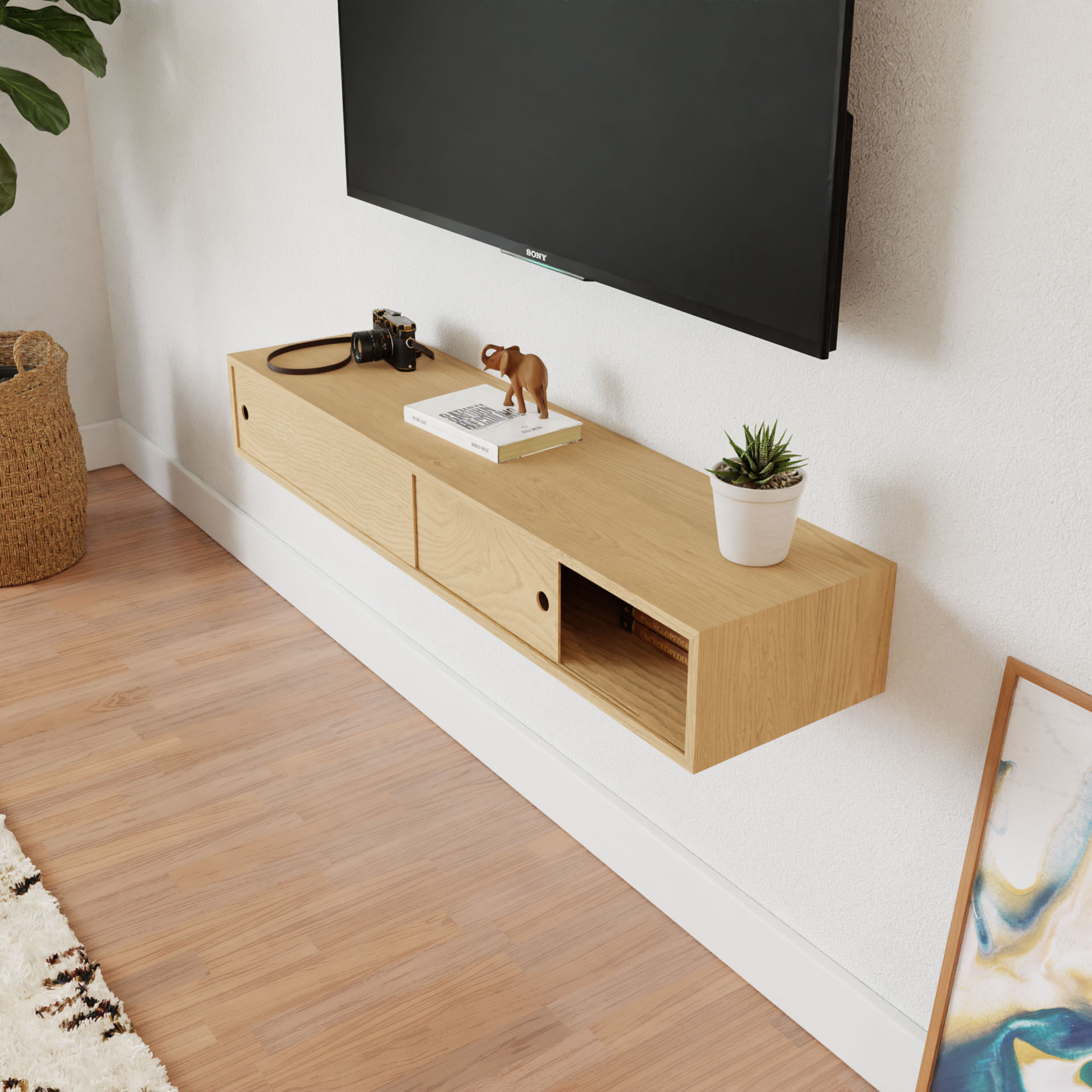 A minimalist living space has a Krovel Media Console Table in Maple mounted beneath a flat-screen TV. The console holds a camera, figurine, and small potted plant. An open drawer reveals its contents while a woven basket and framed artwork rest invitingly on the floor nearby.