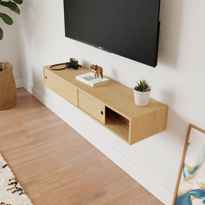 A minimalist living space has a Krovel Media Console Table in Maple mounted beneath a flat-screen TV. The console holds a camera, figurine, and small potted plant. An open drawer reveals its contents while a woven basket and framed artwork rest invitingly on the floor nearby.