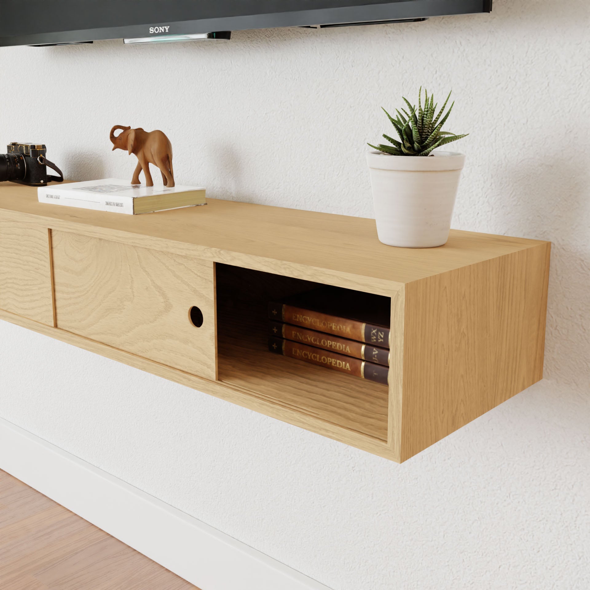 A Krovel Media Console Table in Maple, crafted from American Cherry and finished with VOC-Free Polymerized Linseed Oil, holds a small potted plant, an elephant figurine, a camera, and some books labeled "Encyclopedia." It is mounted on a white wall below a television.