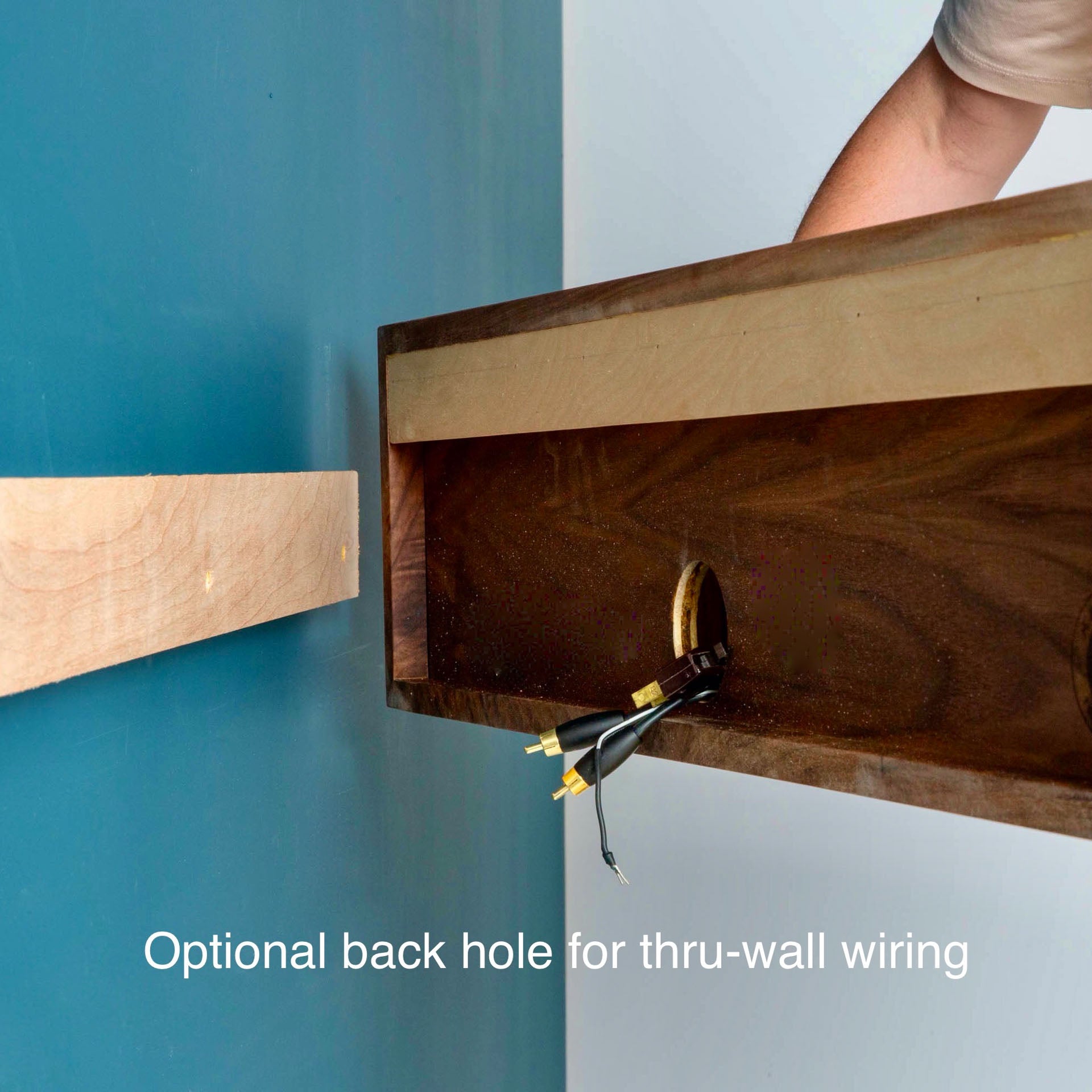 Someone is installing a Krovel Media Console Table in Walnut on a blue wall. The shelf includes an optional back hole for through-wall wiring, coated with VOC-Free Polymerized Linseed Oil, and there are cables emerging from the circular opening.