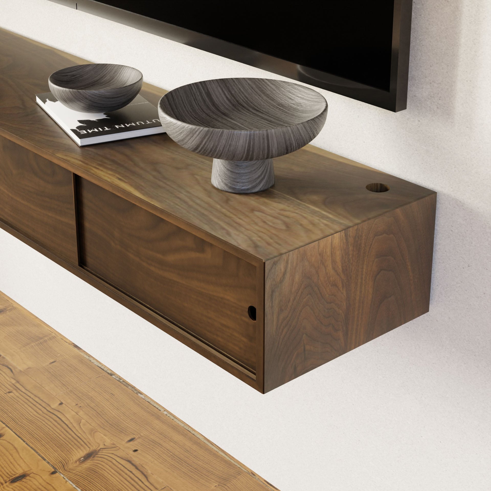 A minimalist interior features a Krovel Media Console Table in Walnut mounted on a white wall. The table displays two modern textured gray bowls of different sizes, with a complementary wooden floor. Part of a flat-screen TV is visible above the console.