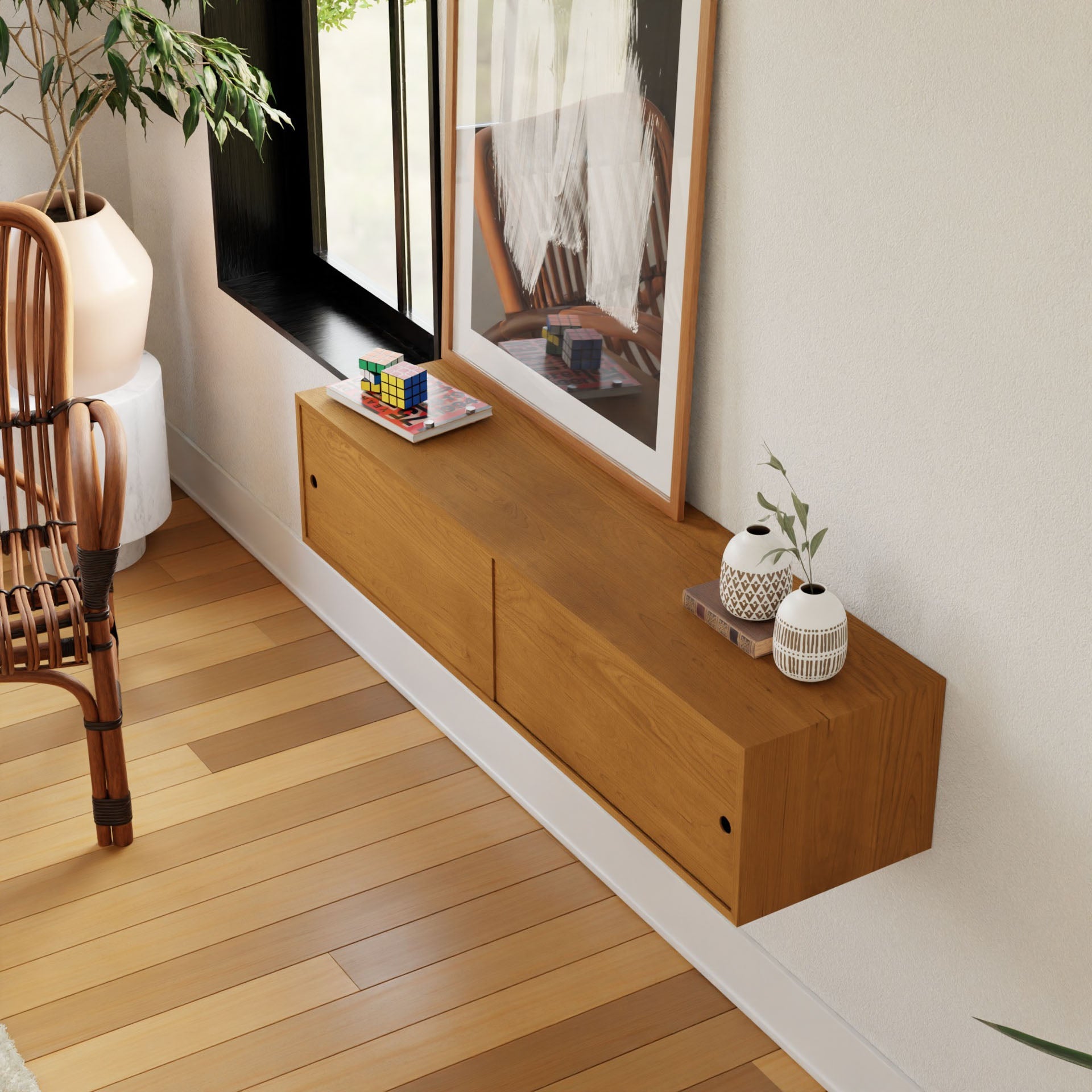 The Krovel Hardwood Oversized Media Console, featuring two drawers, is mounted on a white wall beneath a window. Decorative vases, a Rubik’s cube, and a framed picture sit on top. A chair and wooden flooring complete the scene.