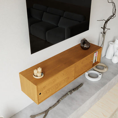 The Krovel Hardwood Oversized Media Console sits beneath a large black TV, styled with candles, a vase, and a book. Nearby on the gray surface are white ceramics, dried branches, and a wooden floor.