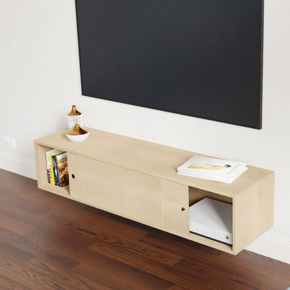 The Krovel Hardwood Oversized Media Console’s sliding doors conceal books, a bowl, and a magazine. Wall mounted under a large black rectangle, it contrasts with the dark wood floor for a modern look.