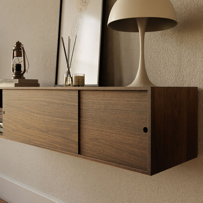 The Krovel Hardwood Oversized Media Console with sliding doors is styled with a beige lamp, framed line drawing, lantern, reed diffusers, and glass cube against a textured beige wall.
