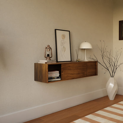 The Krovel Hardwood Oversized Media Console, styled as a modern wall-mounted shelf, displays books, a sketch, a lantern, and decor above a wood floor. A large white vase with branches stands nearby against the beige wall.
