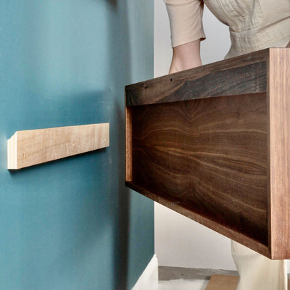 Dressed in beige, the person is skillfully assembling a Krovel Oversized Media Console Table in Walnut against a blue wall, attaching an elegant wood panel onto a light bracket. Their hands and torso are visible as they work on this stylish American Cherry-inspired addition to the room.