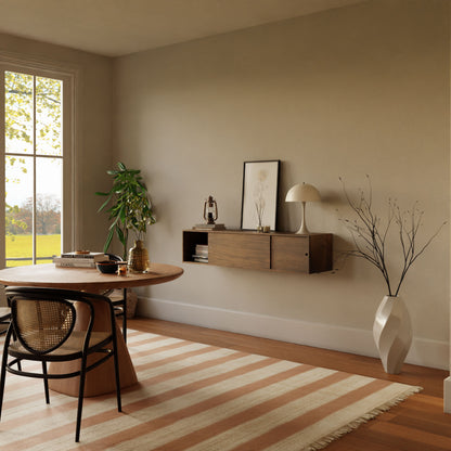A cozy room with a large garden-view window features a round hand-finished American Cherry wood table and a wicker chair on a striped rug. A Krovel Oversized Media Console Table in Walnut holds decor items and a lamp, while a tall vase with branches and potted plant enhance the modern minimalistic setting.