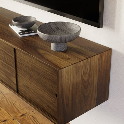 A close-up of the Krovel Hardwood Oversized Media Console styled as a floating unit, with two gray decorative bowls and a magazine on top, set against a white wall and wooden floor.