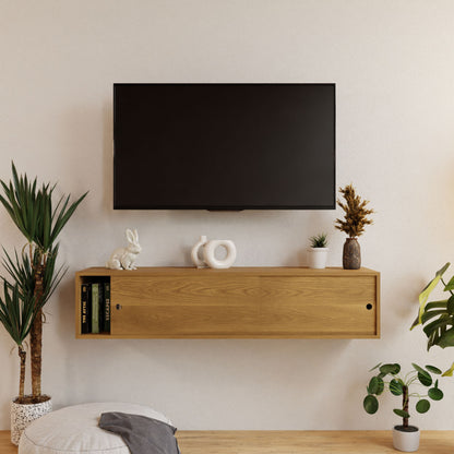 A wall-mounted flat-screen TV is above a Krovel Oversized Media Console Table in White Oak, decorated with items like a white rabbit figurine, stacked books, and a potted plant. A round pouf and an additional floor plant complement the cozy space.