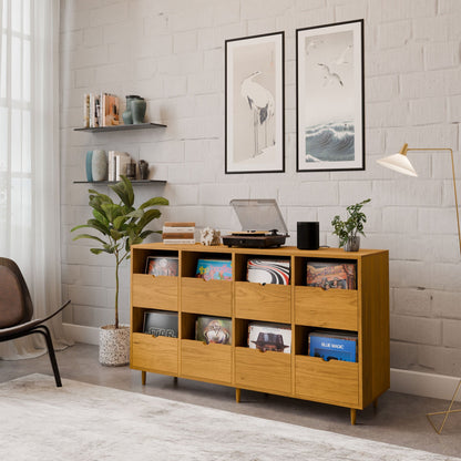A cozy, modern living room features the Krovel Hardwood Eight Drawer Record Credenza with maple drawers for vinyl albums and a turntable. Above are two framed art prints; nearby, shelves hold books, a potted plant, and a floor lamp on a light rug.