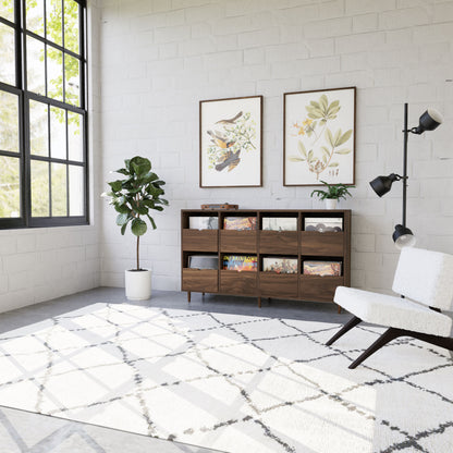 Bright living room with a large window, potted plant, Krovel Hardwood Eight Drawer Record Credenza with books and magazines, two botanical art prints on the wall, white cushioned chair, and a geometric patterned rug.