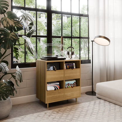 A modern living room showcases a Krovel Hardwood Four Drawer Record Credenza holding a record player, headphones, and vinyl records near large windows. The space features a cozy couch, floor lamp, and lush plant bathed in natural light.