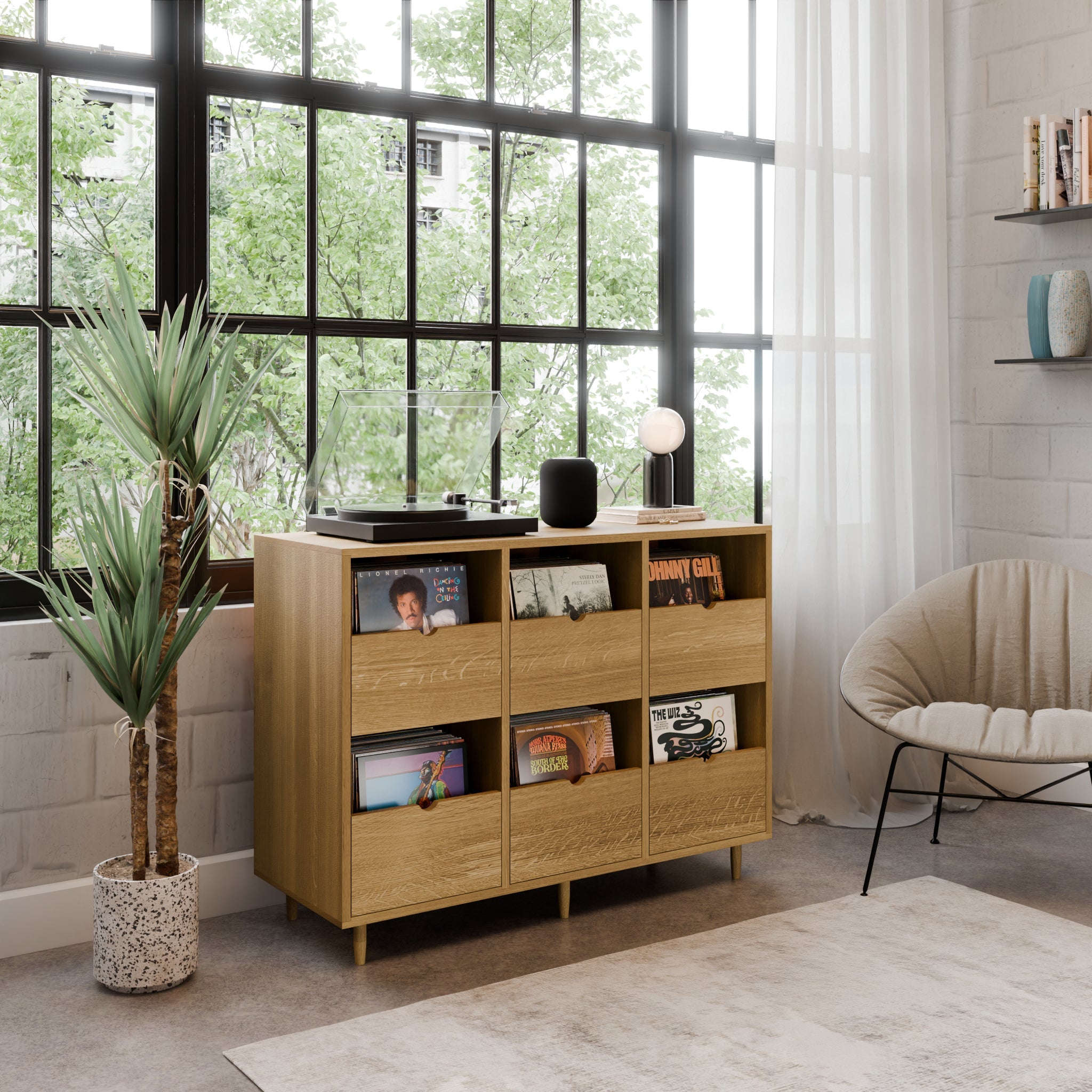 The Krovel Hardwood Six Drawer Record Credenza, handcrafted with maple soft-close drawers, stores vinyl records and a player by large windows. A potted plant, cozy chair, and bookshelves complete the sunlit modern living room.
