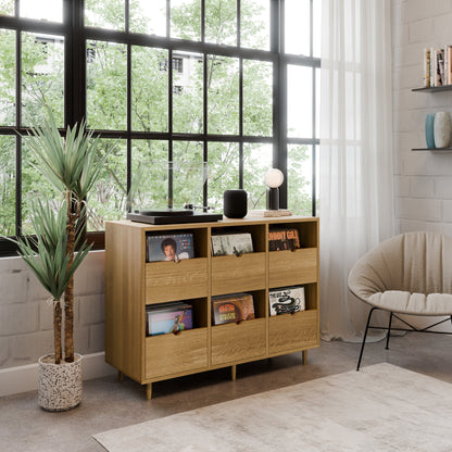 The Krovel Hardwood Six Drawer Record Credenza, handcrafted with maple soft-close drawers, stores vinyl records and a player by large windows. A potted plant, cozy chair, and bookshelves complete the sunlit modern living room.