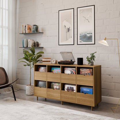 A modern living room with a Krovel Hardwood Eight Drawer Record Credenza, vinyl records, a turntable, a potted plant, bird wall art, bookshelves, and a floor lamp set against a white brick wall.
