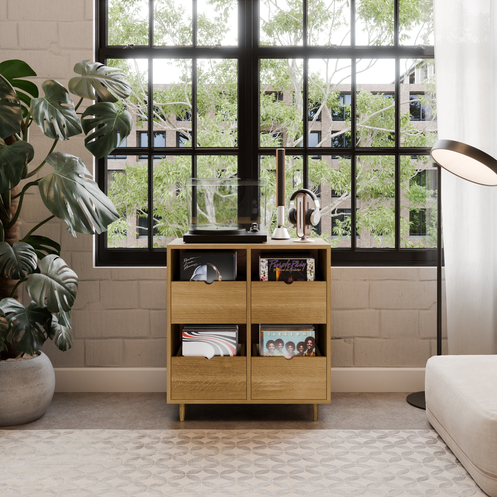 A modern living room corner features the Krovel Hardwood Four Drawer Record Credenza with maple drawers for vinyls, a record player, and headphones. Set by a black-framed window with green views, it’s accented by a floor lamp and lush leafy plant.