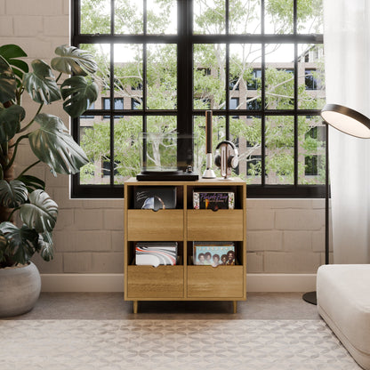 A modern living room corner features the Krovel Hardwood Four Drawer Record Credenza with maple drawers for vinyls, a record player, and headphones. Set by a black-framed window with green views, it’s accented by a floor lamp and lush leafy plant.