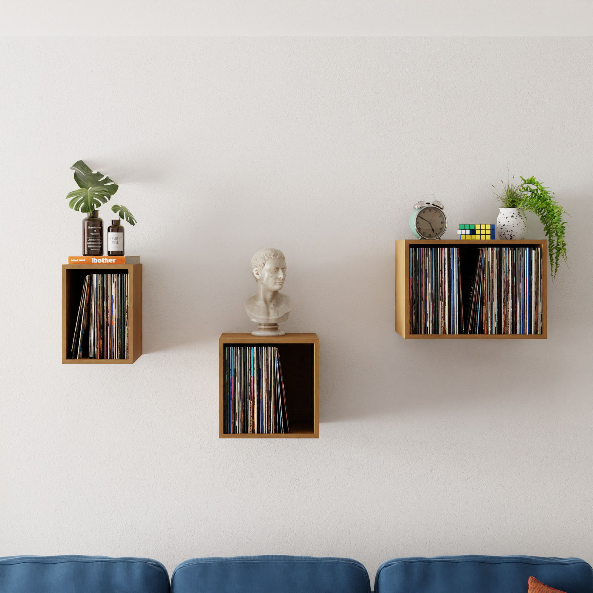 Three Record Storage Shelves in Cherry from Krovel hold vinyl records, a small plant, books, a bust, and an alarm clock above a blue sofa against a cream-colored wall, adding mid-century modern elegance.
