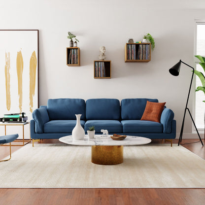 A modern living room showcases a blue sofa with an orange cushion, complemented by a white marble coffee table on a beige rug. Krovel's Record Storage Shelves in Cherry enhance the decor, while an abstract painting and floor lamp complete this mid-century modern aesthetic.