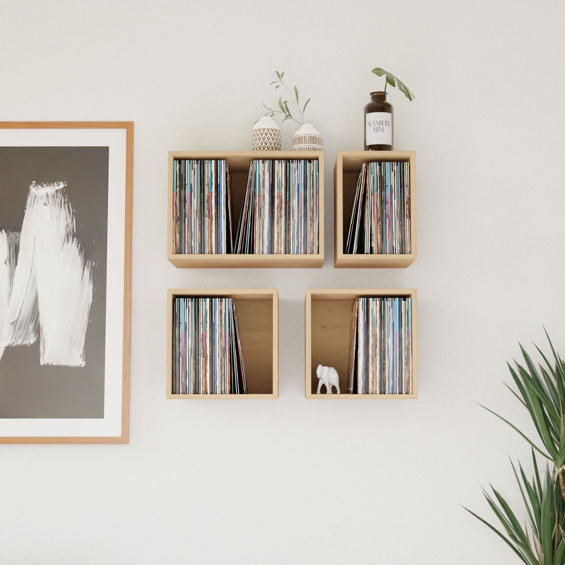 Four Krovel Maple record shelves hold vinyl records. Two small vases with plants and an "Amber Musk" jar sit on top. An abstract painting in a wooden frame hangs to the left, with green plant leaves visible at the bottom right.