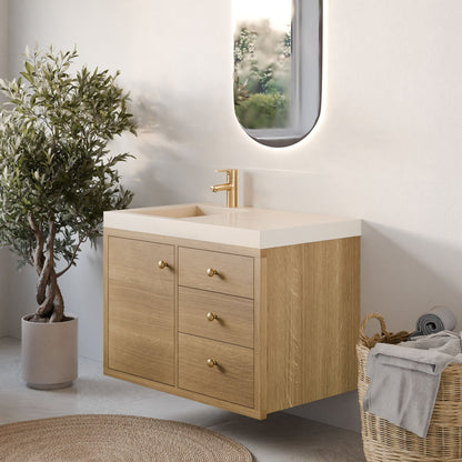 A modern bathroom features the Krovel Furniture Co. Maine Made Single Door, Triple Drawer Vanity with a gold faucet and oval backlit mirror. A potted plant and a wicker basket with towels enhance the serene, neutral space filled with natural light.