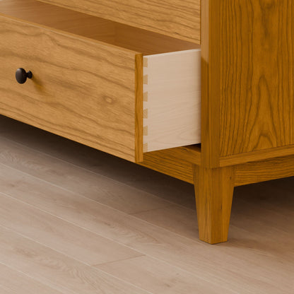 The Six Drawer Dresser in Cherry by Krovel Furniture Co. stands on a light wooden floor, crafted from solid hardwood with a natural wood grain finish and simple black knobs. One drawer is partially open, revealing its smooth, light-colored interior—a testament to sustainable construction.