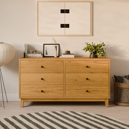 The Six Drawer Dresser in Cherry by Krovel Furniture Co. stands against the wall, topped with decor items, books, and plants. A modern geometric artwork hangs above it. Nearby are a round floor lamp and a woven basket on a light floor partly covered by a striped rug.