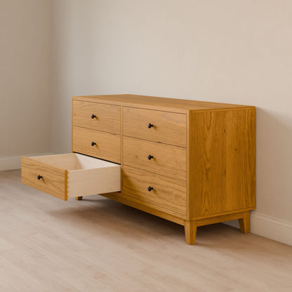 The Maine Made Six Drawer Dresser by Krovel Furniture Co. stands against a light wall on a light wood floor, with one left drawer open and the others closed. It features solid wood veneer and is handmade in Portland, Maine.