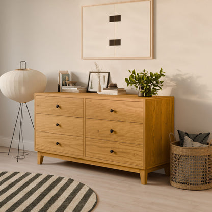 A Krovel Furniture Co. Six Drawer Dresser in Cherry, featuring black knobs and sustainable construction, stands adorned with books, a potted plant, and decor. A framed abstract art piece hangs above while a basket, floor lamp, and round striped rug complete the light hardwood flooring setting.