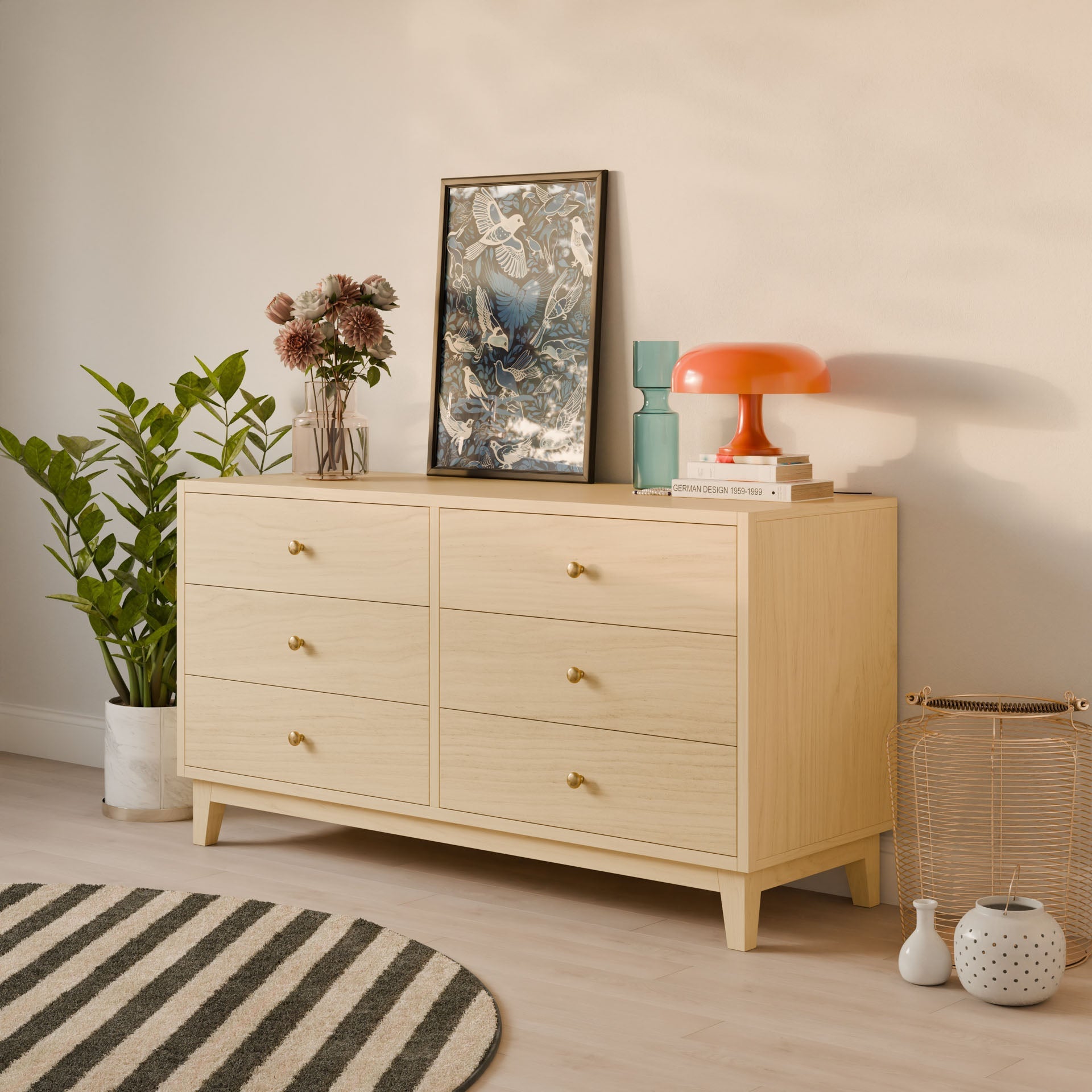 The Six Drawer Dresser in Maple from Krovel Furniture Co. showcases framed artwork, books, and an orange lamp on top. It stands on a wooden floor with a striped rug, accompanied by a potted plant and decorative basket. Customize its size to fit your space perfectly.