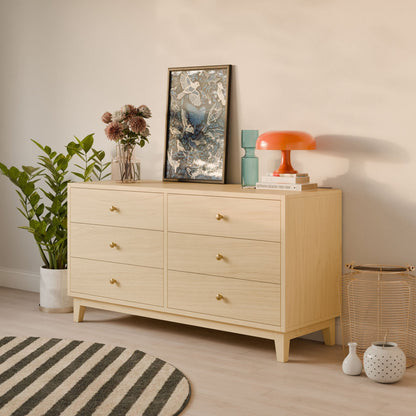 The Six Drawer Dresser in Maple from Krovel Furniture Co. showcases framed artwork, books, and an orange lamp on top. It stands on a wooden floor with a striped rug, accompanied by a potted plant and decorative basket. Customize its size to fit your space perfectly.