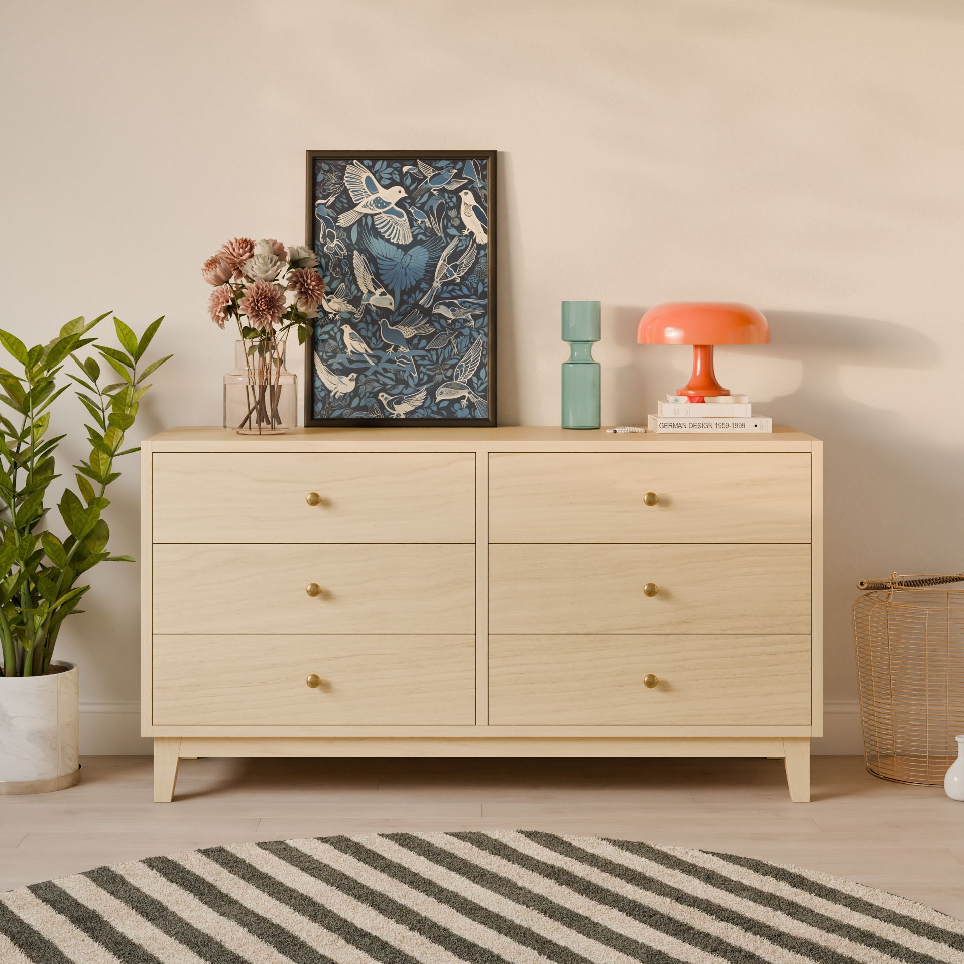 The Six Drawer Dresser in Maple by Krovel Furniture Co. features gold knobs and supports a framed floral artwork, a green flower-filled vase, and an orange lamp. Crafted from eco-friendly premium plywood, it stands next to a plant on the left, set on a striped rug.