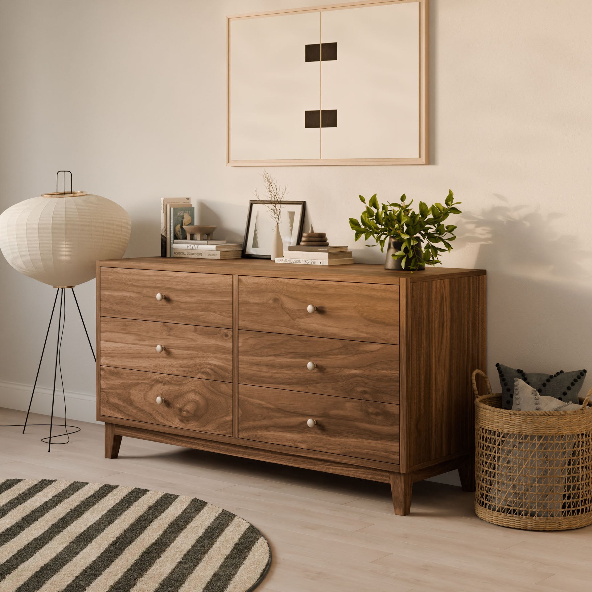 A contemporary living room highlights the Krovel Furniture Co. Six Drawer Dresser in Walnut, featuring a solid hardwood face frame. Books, a plant, and decor rest atop it, with minimalist artwork above. A woven basket with pillows sits beside it, complemented by a round floor lamp and striped rug.