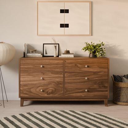 The Krovel Furniture Co.s Six Drawer Dresser in Walnut, crafted from solid hardwood face frames and premium cabinet grade plywood, displays books, a plant, and a vase. An abstract art piece is above; a large floor lamp is on the left, with a woven basket atop a striped rug to the right.