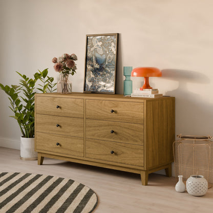 The Krovel Furniture Co. Six Drawer Dresser in White Oak holds a framed artwork, plant, teal vase, stacked books, and an orange lamp. A tall leafy plant is on the left; a woven basket on the right. Black and white striped rug completes the scene.