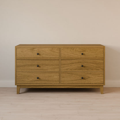 The Six Drawer Dresser in White Oak by Krovel Furniture Co. features solid hardwood construction with round knobs, set against a light wall and wooden floor. It boasts a simple, modern design with a natural wood finish, ensuring both elegance and durability.