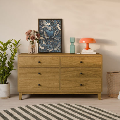 The Krovel Furniture Co. Six Drawer Dresser in White Oak, crafted from formaldehyde-free plywood, is topped with framed artwork, a pink lamp, and books. With a potted plant on the left and a wicker basket on the right, it stands on a light wood floor with a striped rug in front.