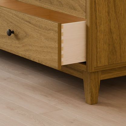 A close-up of a wooden drawer from the Six Drawer Dresser in White Oak by Krovel Furniture Co. highlights dovetail joints crafted from solid hardwood, with a natural wood grain finish and black round knob on light flooring, exemplifying sustainable construction practices.